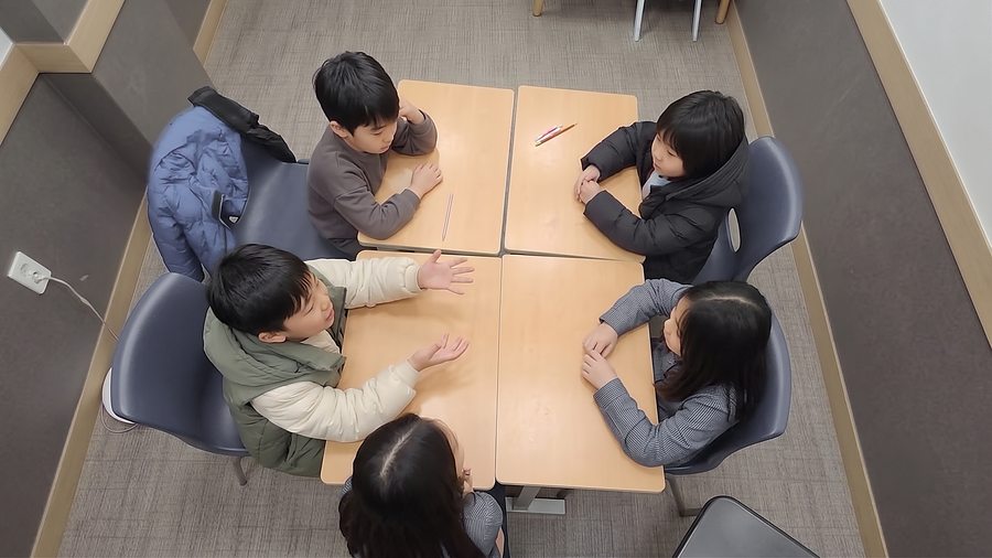 책상에 둘러앉아 한 사람의 이야기를 듣는 아이들